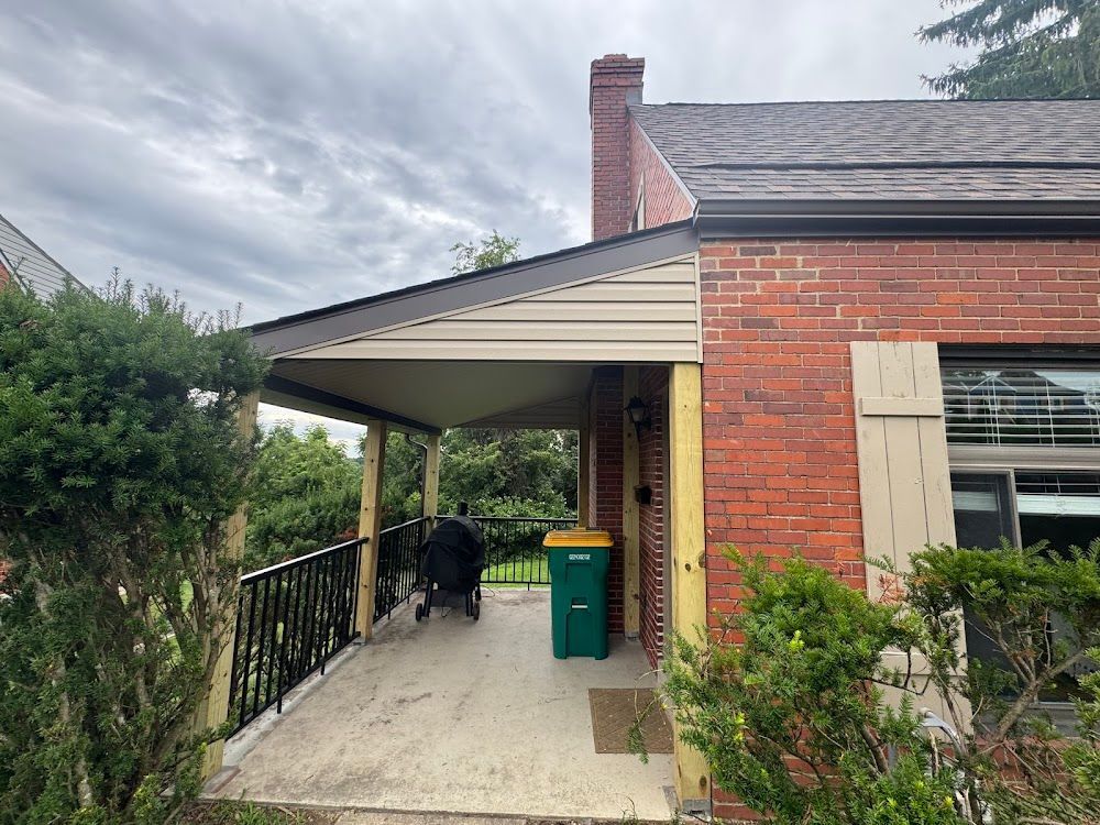 Covered porch attached to a red brick house with a black railing, a green trash bin, and a grill.