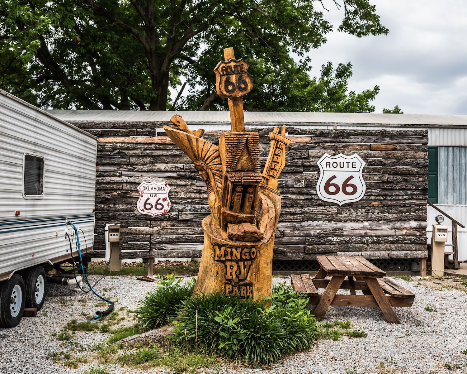 A wooden statue of a totem pole with a route 66 sign on it is in front of a trailer.