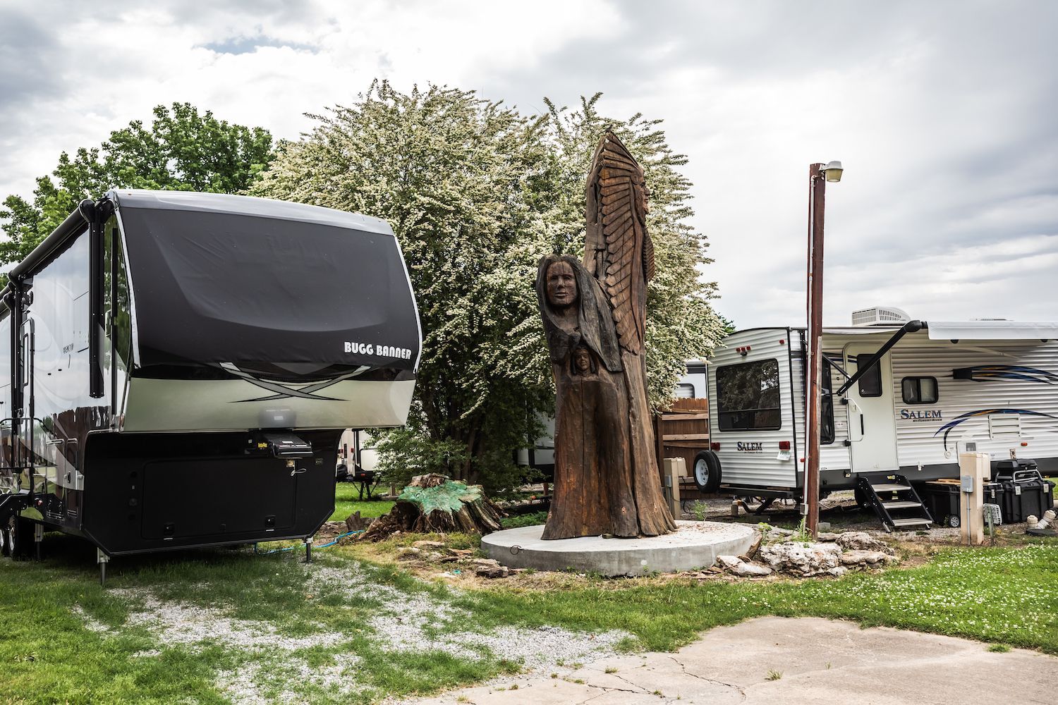 A statue of a woman is sitting in front of a rv parked in a grassy area.
