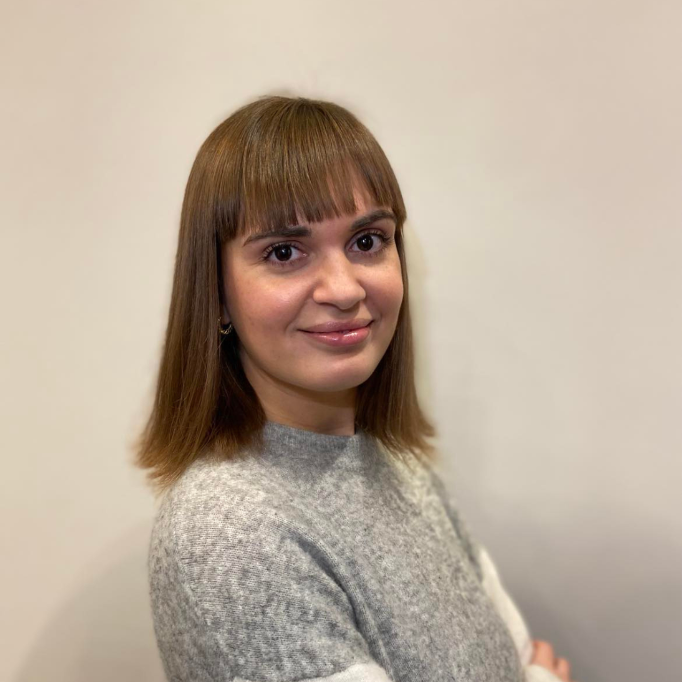 Woman with blonde hair and bangs, wearing a light-colored blazer and gray top, smiling at the camera.