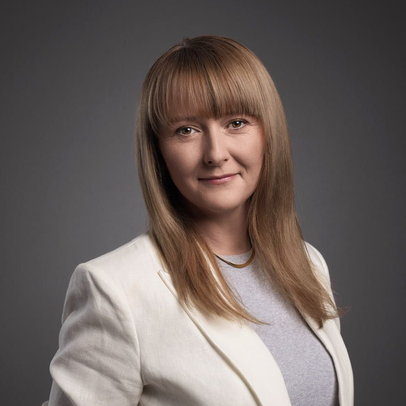 Woman with blonde hair and bangs, wearing a light-colored blazer and gray top, smiling at the camera.