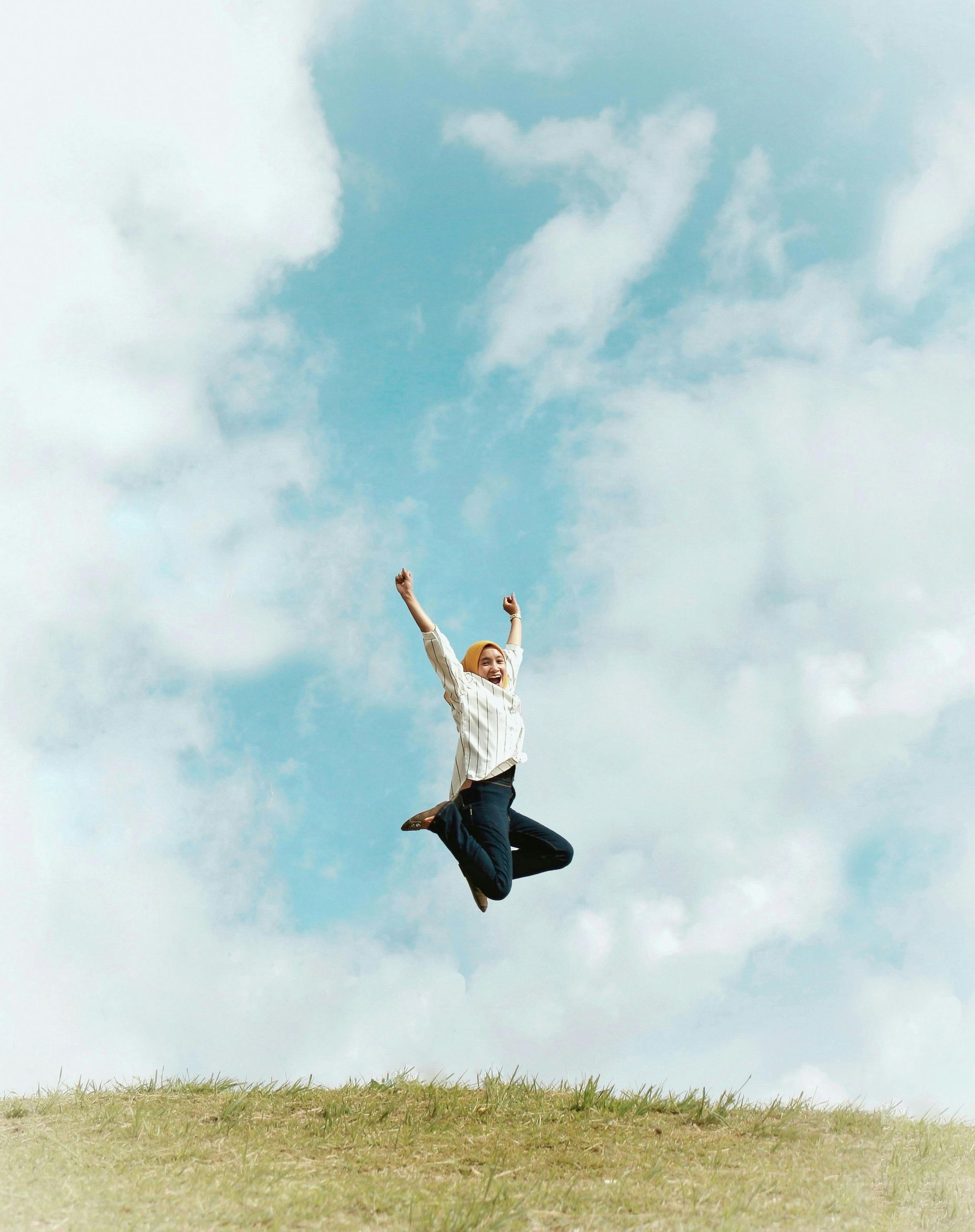 Person jumping joyfully with arms raised against a blue sky with clouds.