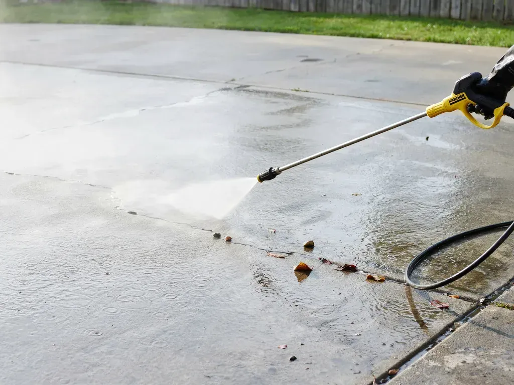 Person pressure-washing a wet driveway with a yellow spray wand, water spraying across the concrete.
