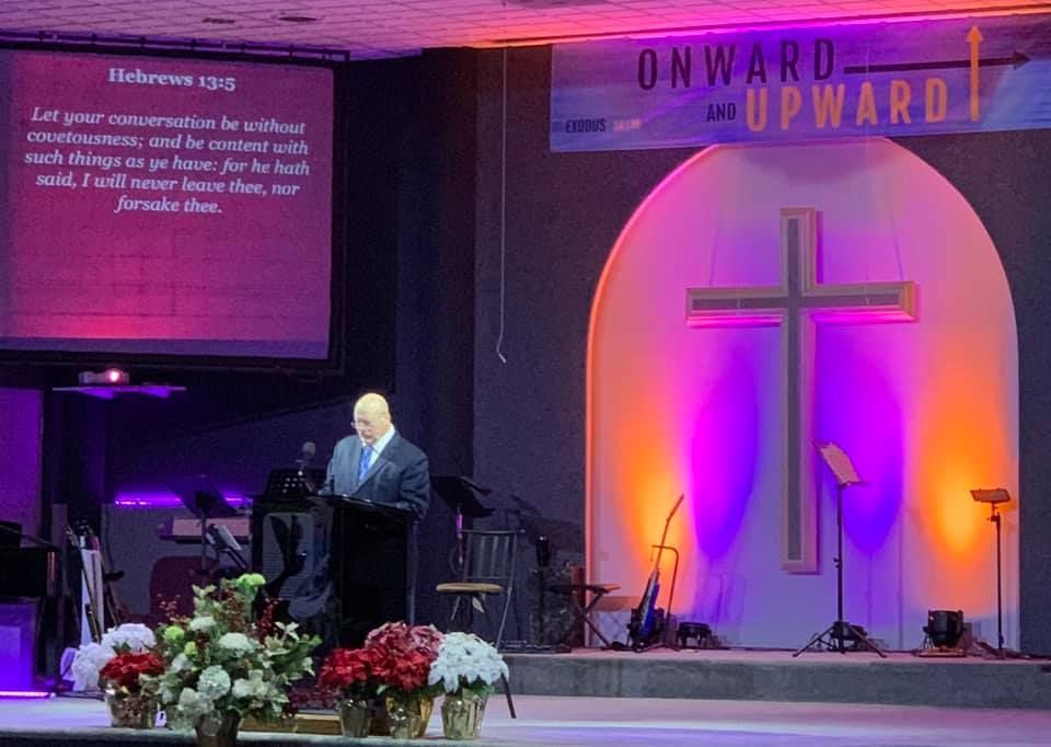 Church sanctuary with pastor at podium, projection screen, cross-lit backdrop, and floral arrangements
