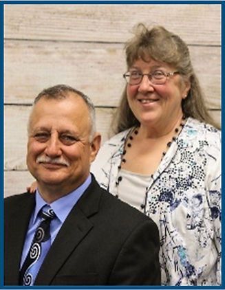 Smiling couple posing indoors against a light wood wall, man in suit and woman in patterned blouse