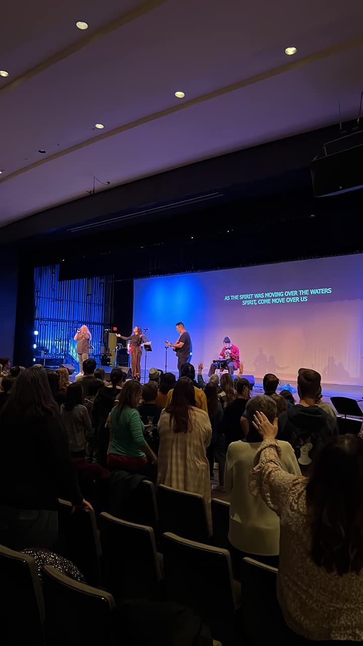 Auditorium worship service with a band on a blue-lit stage and congregants standing and raising hands