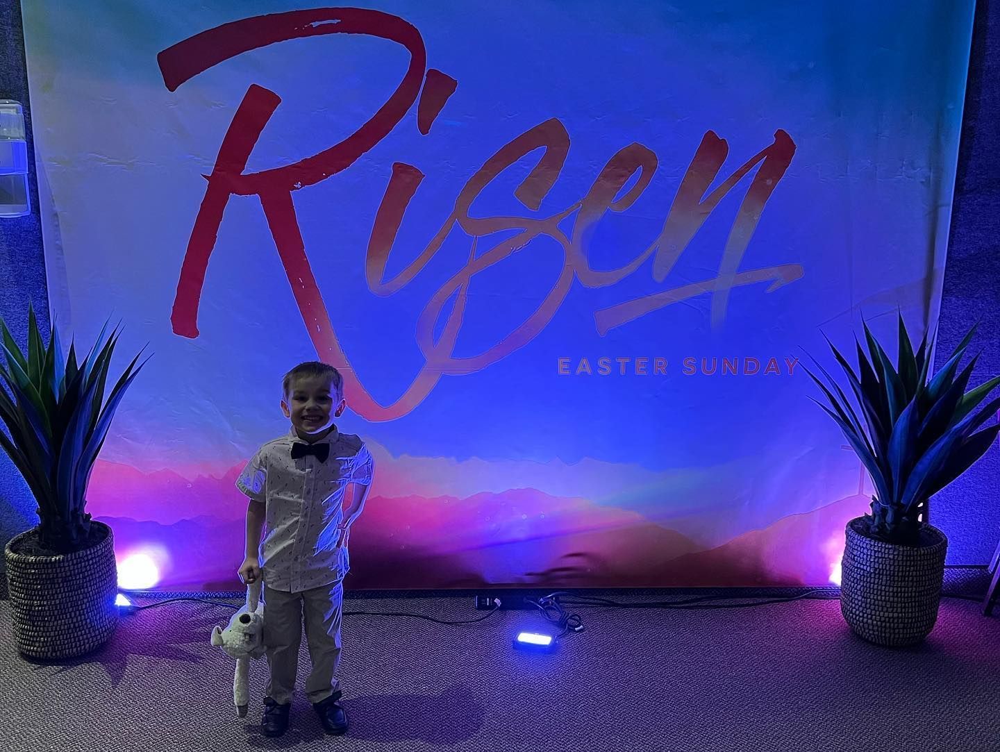 Child on stage in front of a large “Risen” backdrop, flanked by potted plants and purple lighting.
