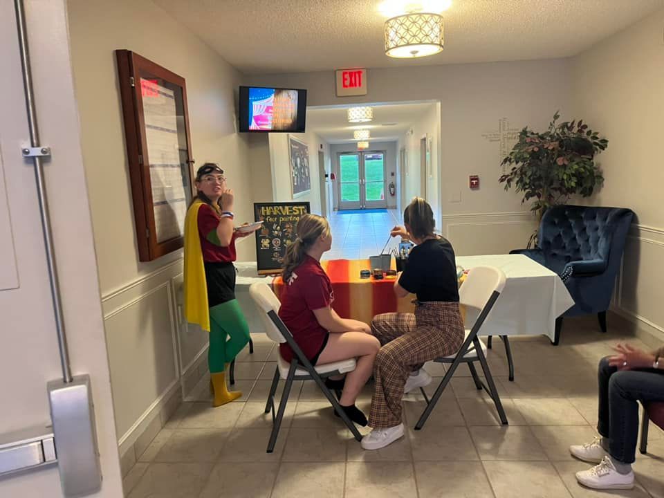 Person presenting to seated attendees in a lobby with a white table, TV, and exit sign.