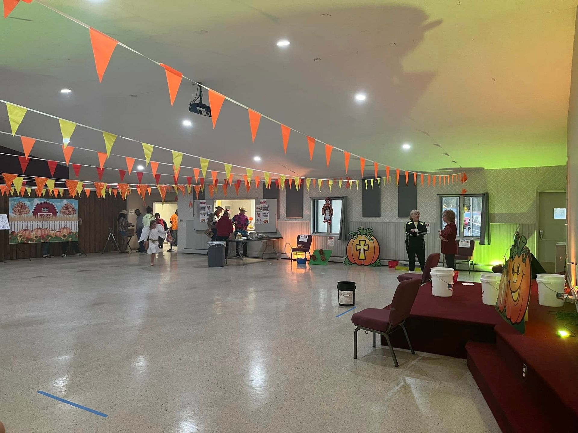 Spacious indoor hall decorated with orange bunting, with people near a stage and tables under green lighting