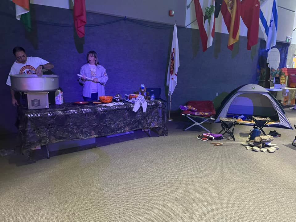 Indoor event with two presenters at a table beside a small tent and equipment, under hanging flags.