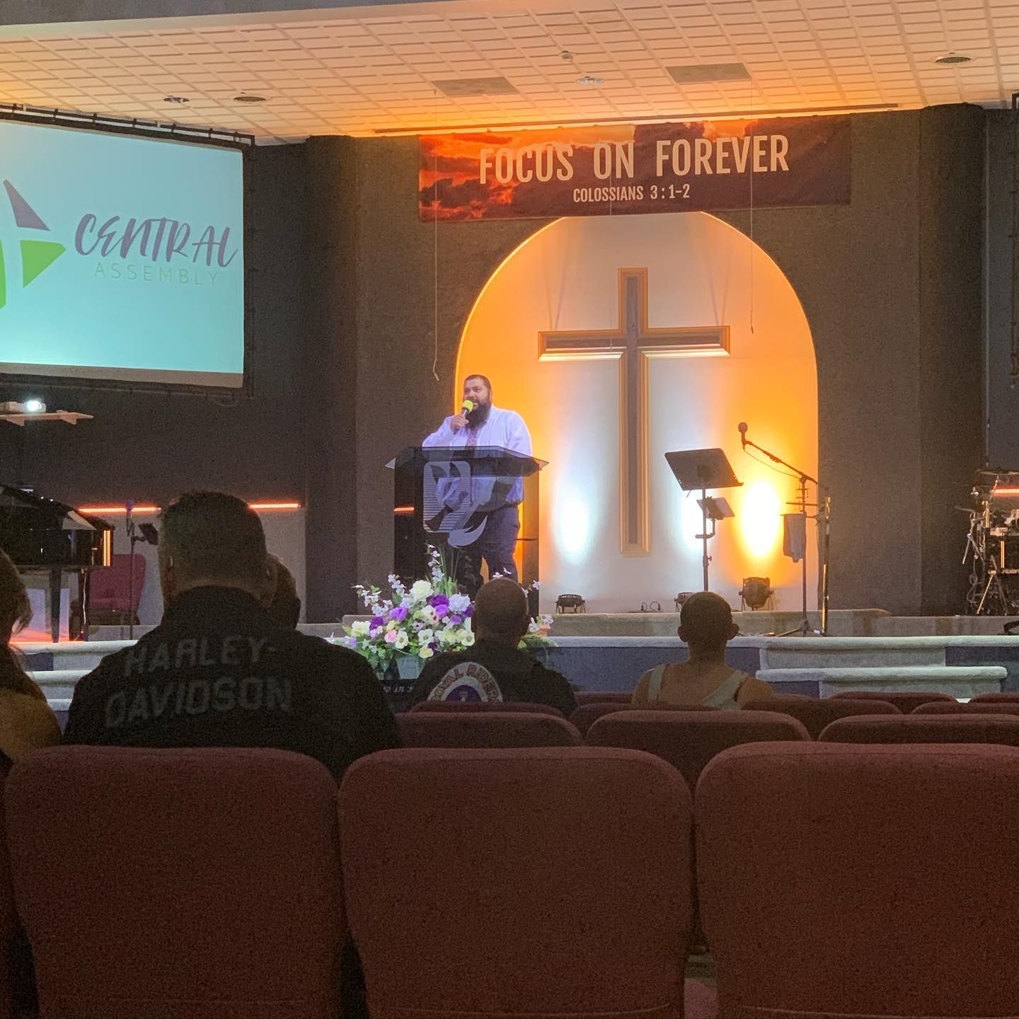 Church stage with speaker at podium, cross lit in warm orange, and banner reading “Focus on Forever”