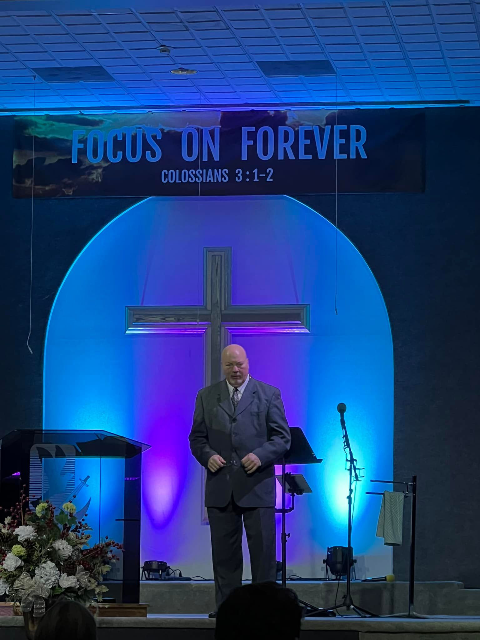 Man standing on a church stage under a blue-purple lit cross with “Focus on Forever” banner above