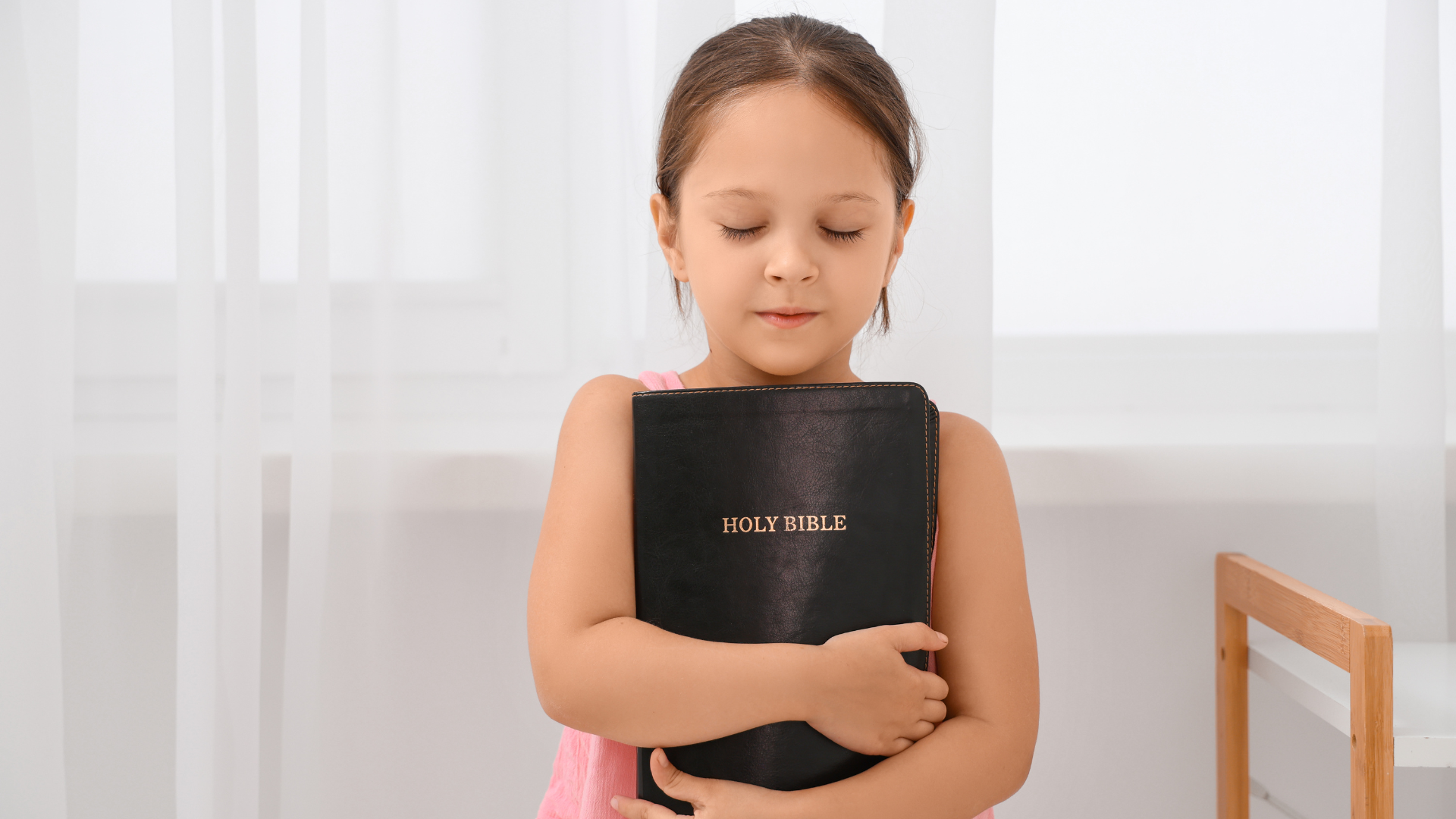 Young girl holding a black notebook against her chest in a bright room