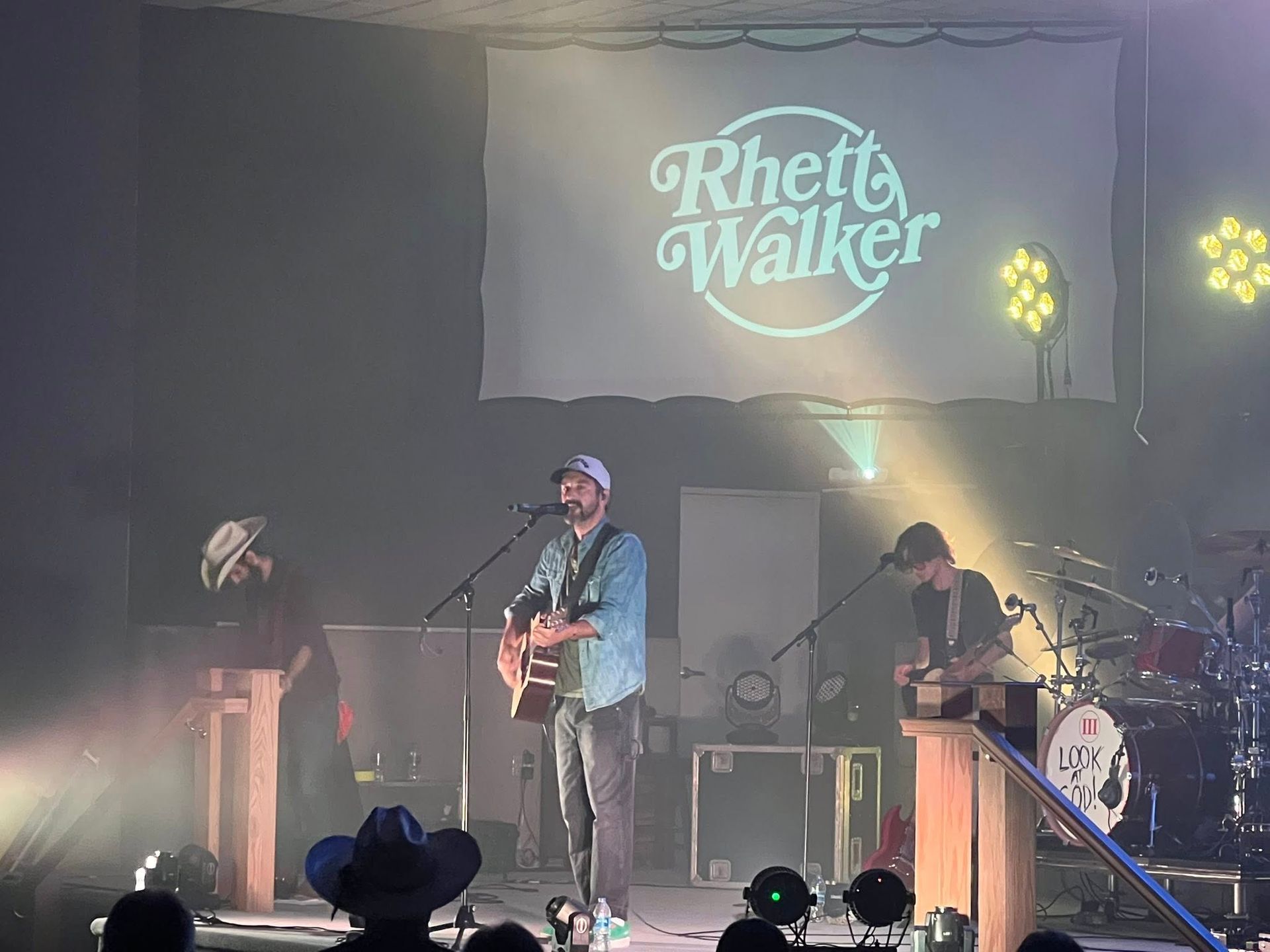 Live concert stage with singer-guitarist under a screen reading “Rhett Walker” and yellow lights