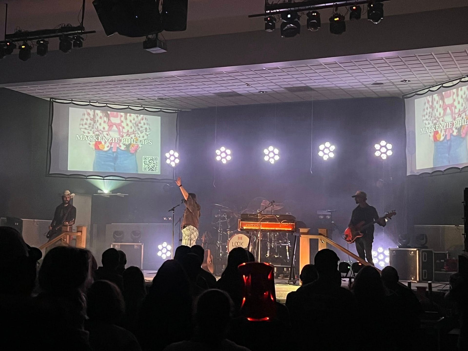 Concert stage with band performing under bright lights, silhouetted audience in the foreground, and projection screens above