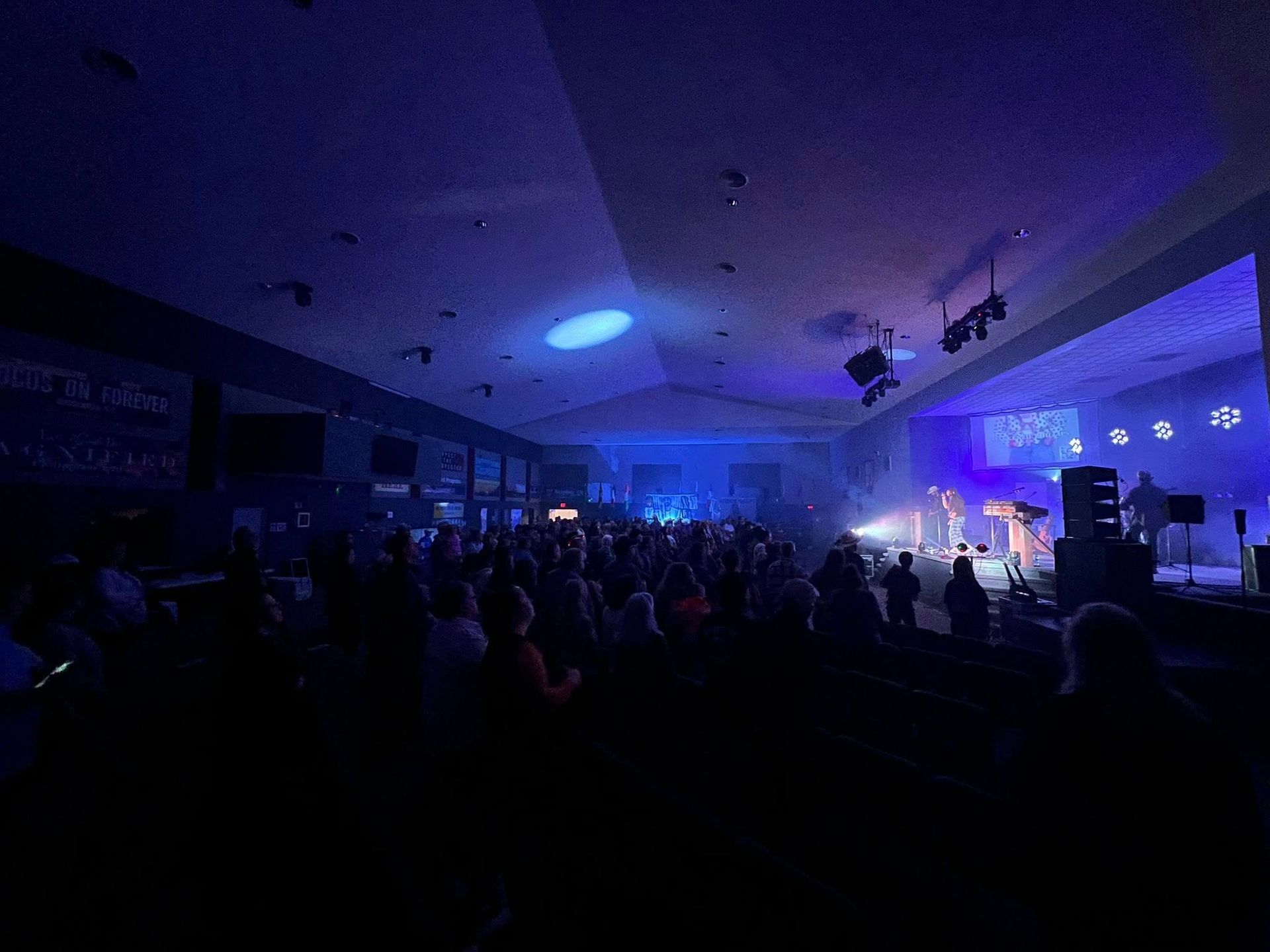 Crowded indoor concert hall lit in blue and purple, with a stage and audience in silhouette