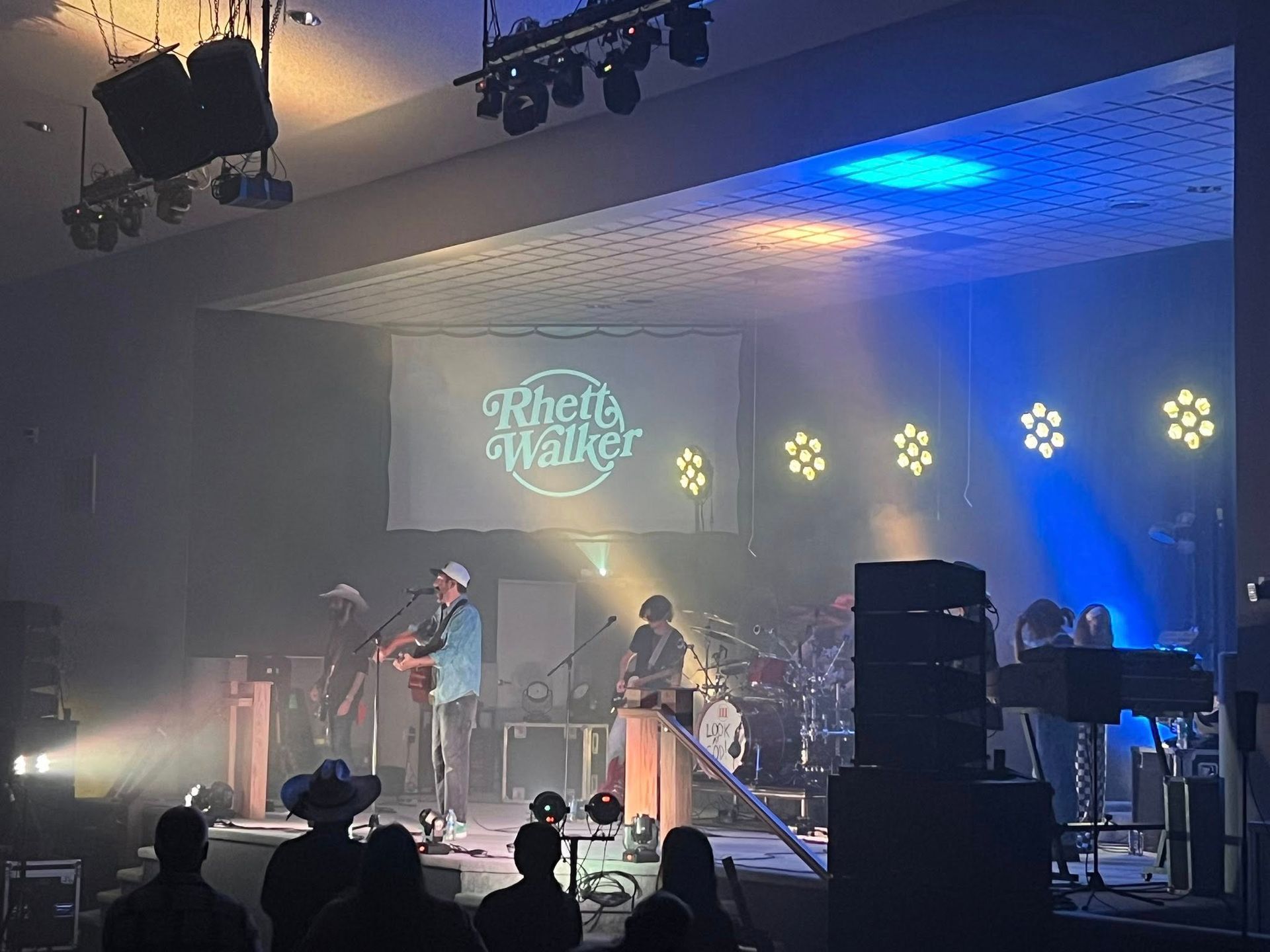 Live band performing on a dimly lit stage with blue and yellow lights and a projected logo backdrop