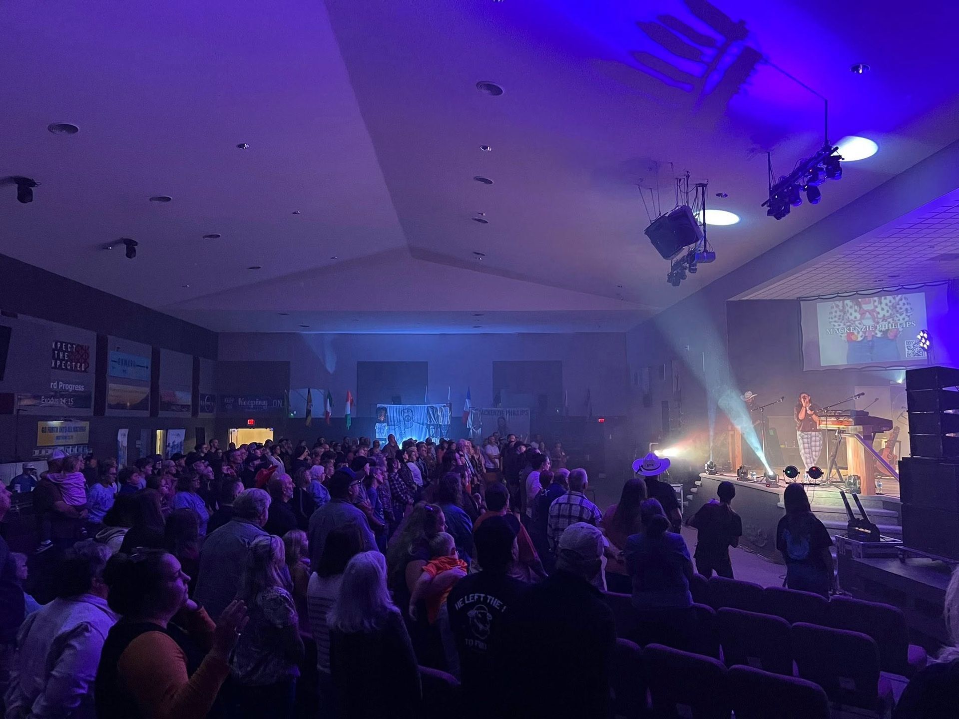 Crowded concert hall with purple lighting, audience facing a brightly lit stage