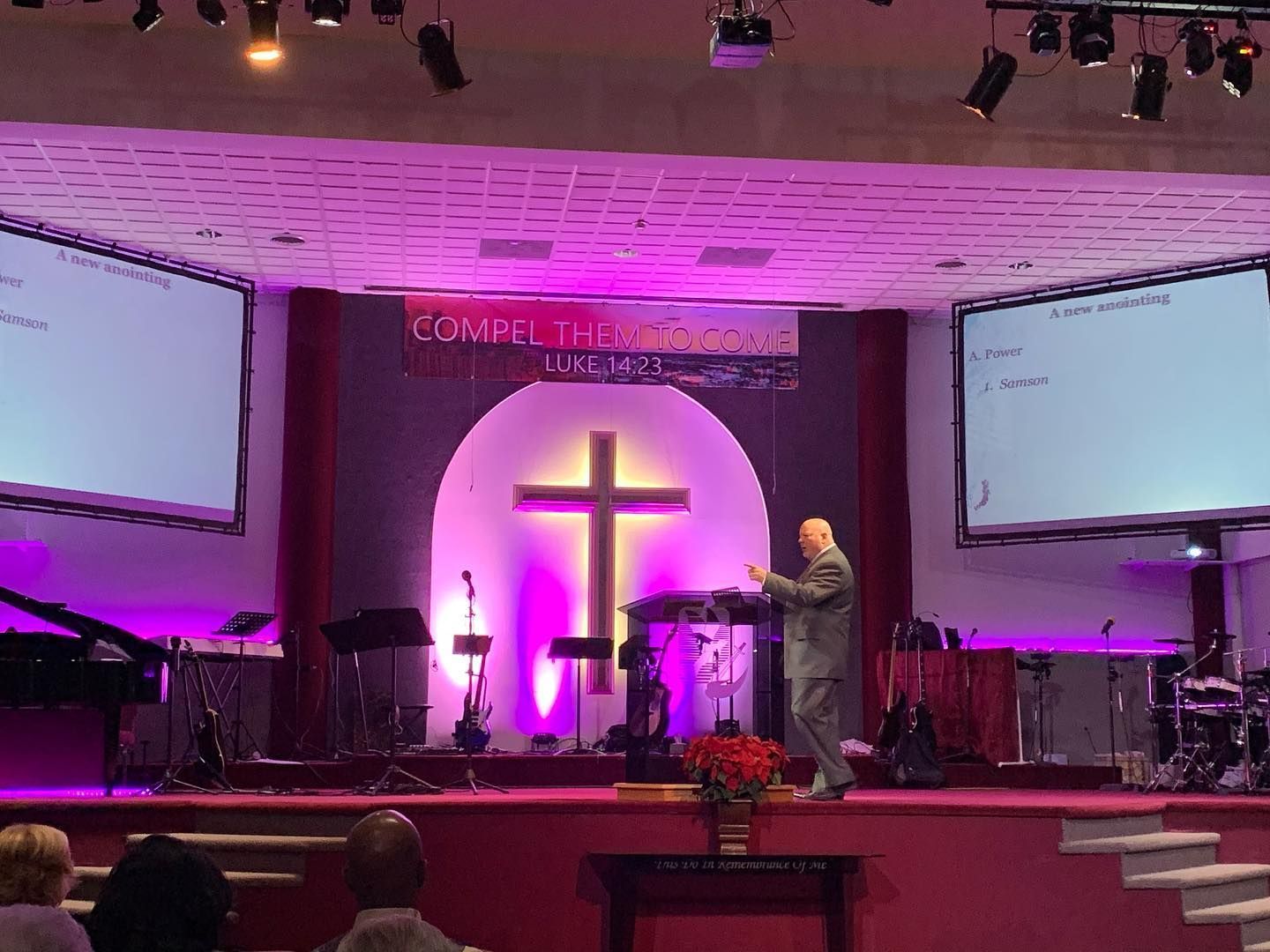 Church stage with illuminated cross, speaker at podium, and projection screens in a purple-lit sanctuary
