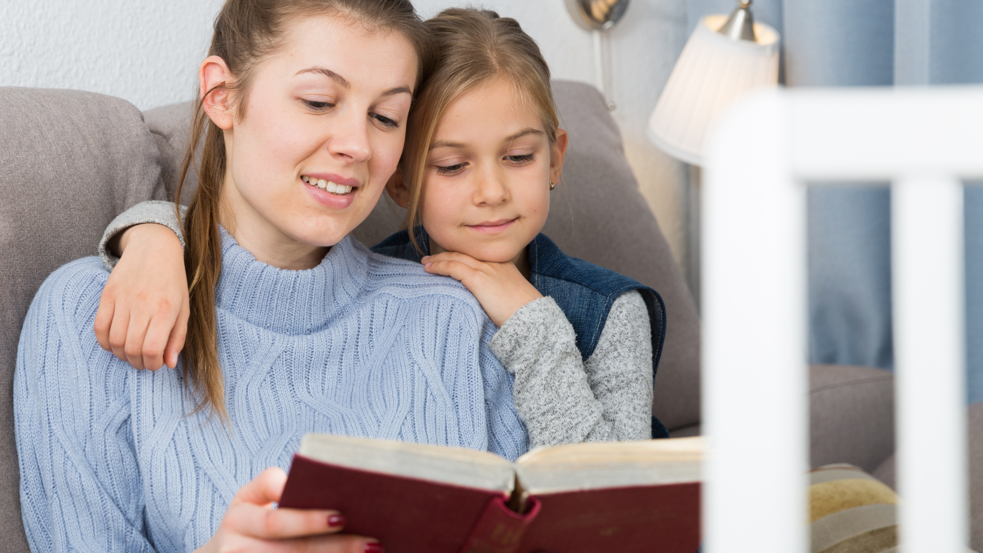 Woman and child reading together on a couch, sharing a red book in a bright living room