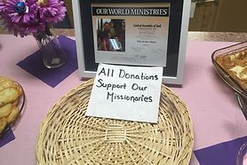 Donation table with sign reading “All Donations Support Our Missionaries” on a purple tablecloth