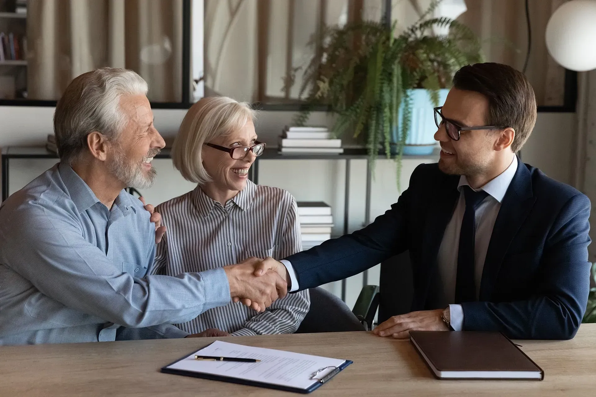 A professional in a suit shakes hands with a person across a table in an office, while another person watches and smiles.