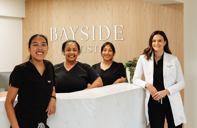 Smiling woman at beach, taking selfie, arms outstretched. Wearing floral top, sunlit, ocean background.