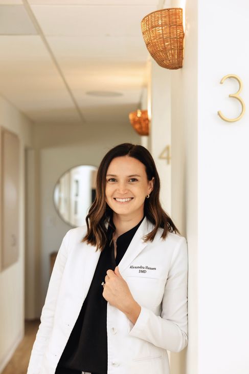 Female dentist in lab coat, smiling, arms crossed, in a dental office hallway.