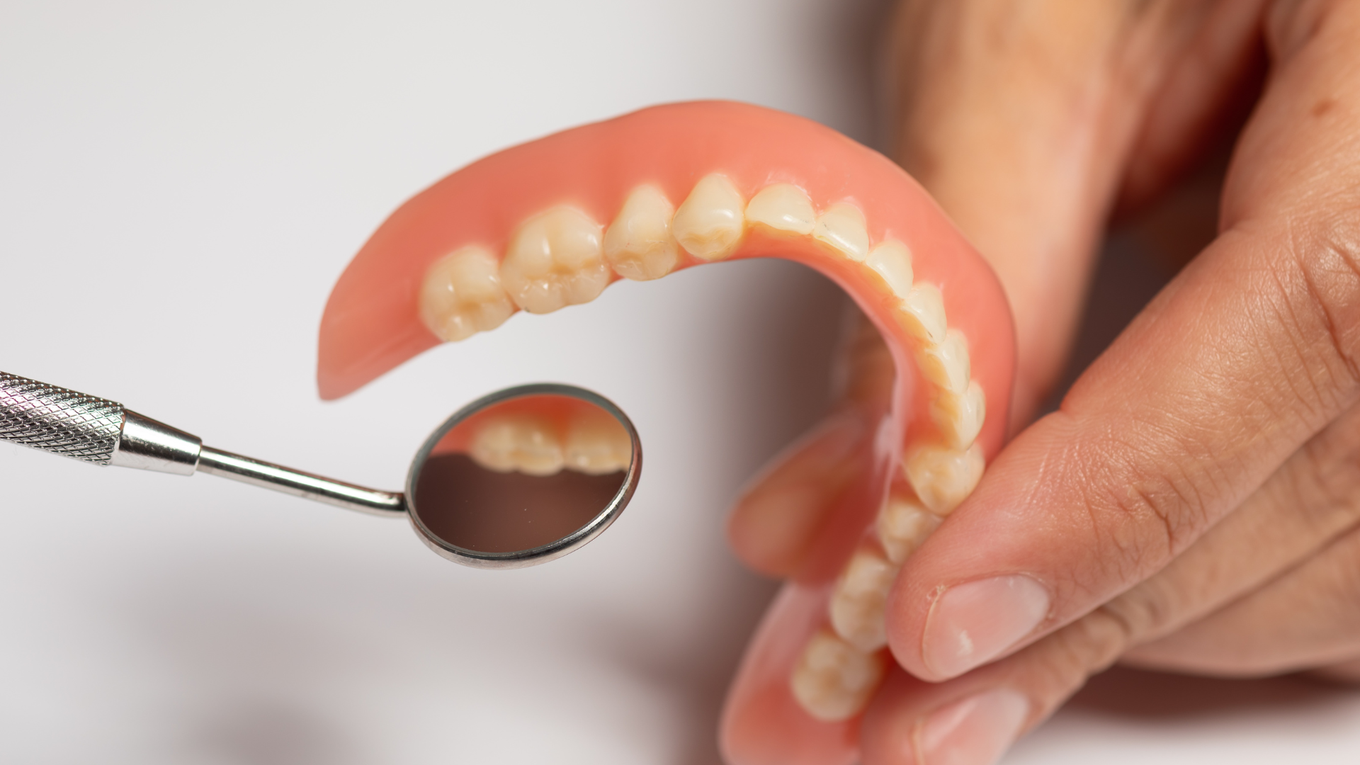 Hand holding a set of dentures and dental mirror. The pink gums and teeth are visible, reflected in the mirror.
