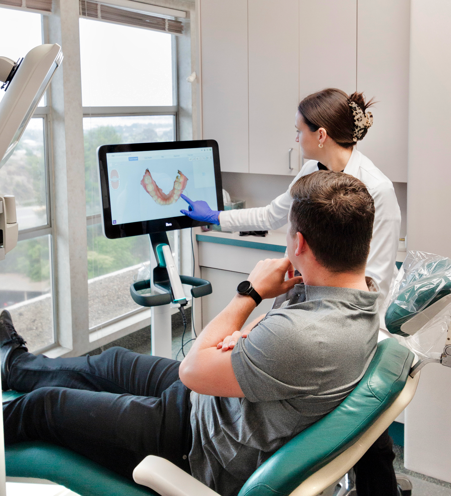 Dentist pointing at teeth scan on screen, explaining to patient reclining in chair, dental office.