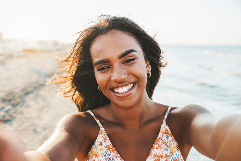 Smiling woman at beach, taking selfie, arms outstretched. Wearing floral top, sunlit, ocean background.