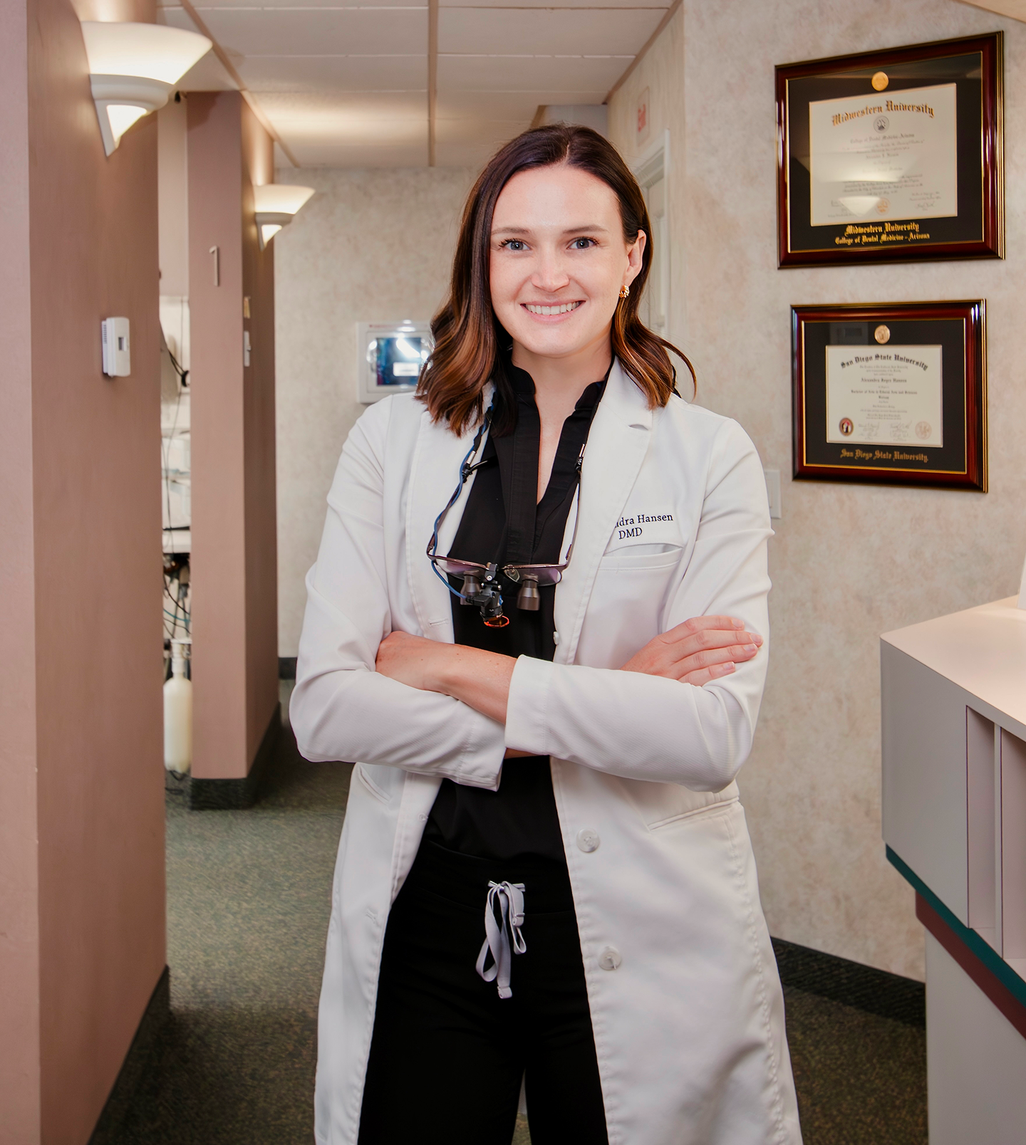 Female dentist in lab coat, smiling, arms crossed, in a dental office hallway.