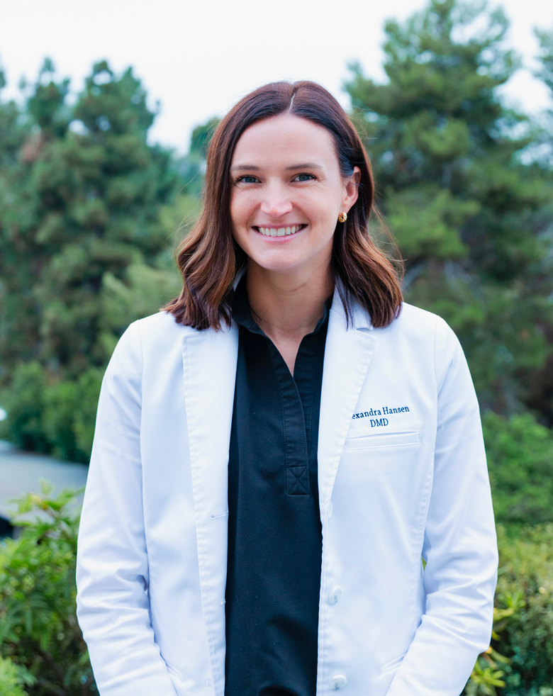 Woman in a white lab coat smiles outdoors; trees in the background.