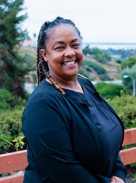 Smiling Black woman with braided hair, wearing black jacket, in outdoor setting.
