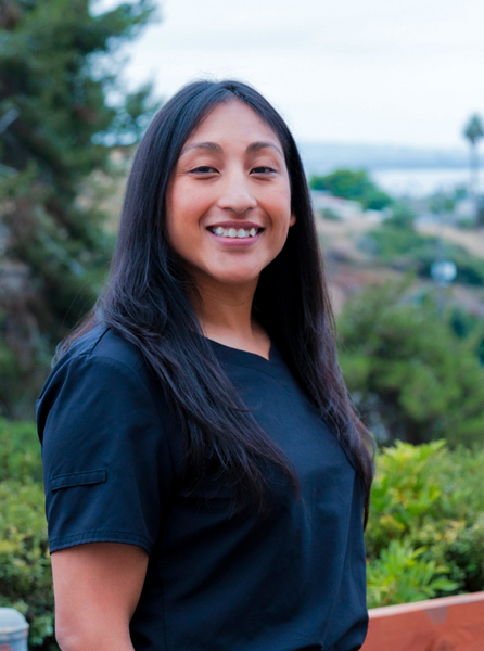 Woman with long dark hair smiles outdoors, wearing a black top, overlooking greenery and water.