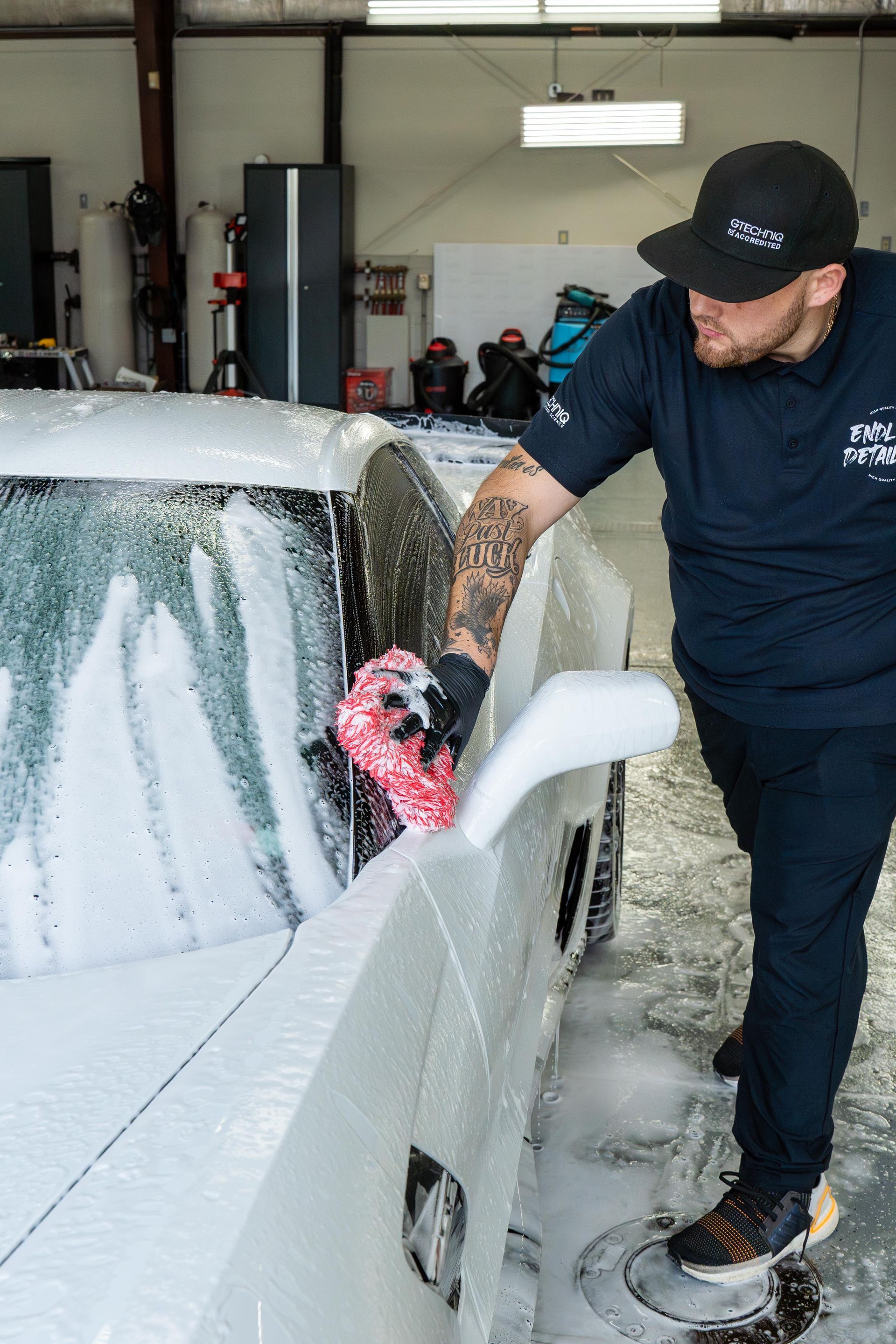 A technician in a black uniform uses a red wash mitt to clean a white car covered in thick soap suds in a garage.