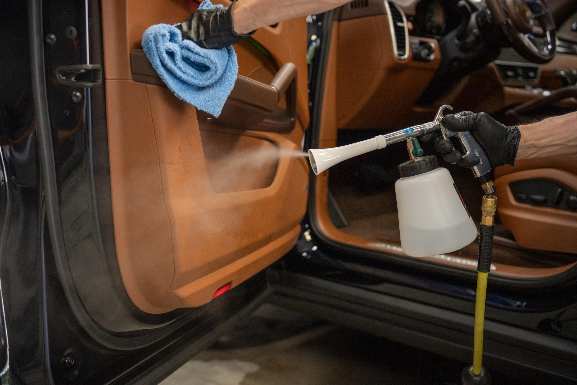 A person in black gloves uses a pneumatic cleaning tool to spray a brown car door interior, holding a blue cloth nearby.