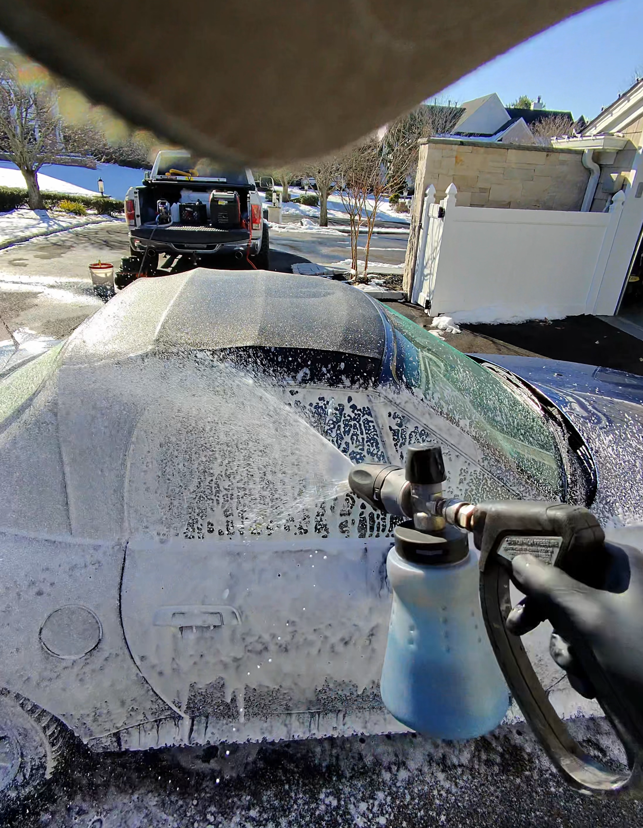 A first-person view of a hand in a black glove using a foam cannon to spray soap onto a car covered in white foam.