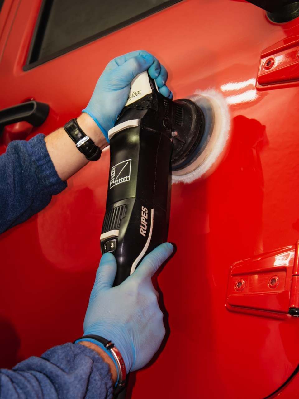 A detailer in a black uniform and cap uses a foam-covered mitt to wash a soap-covered car in a garage.