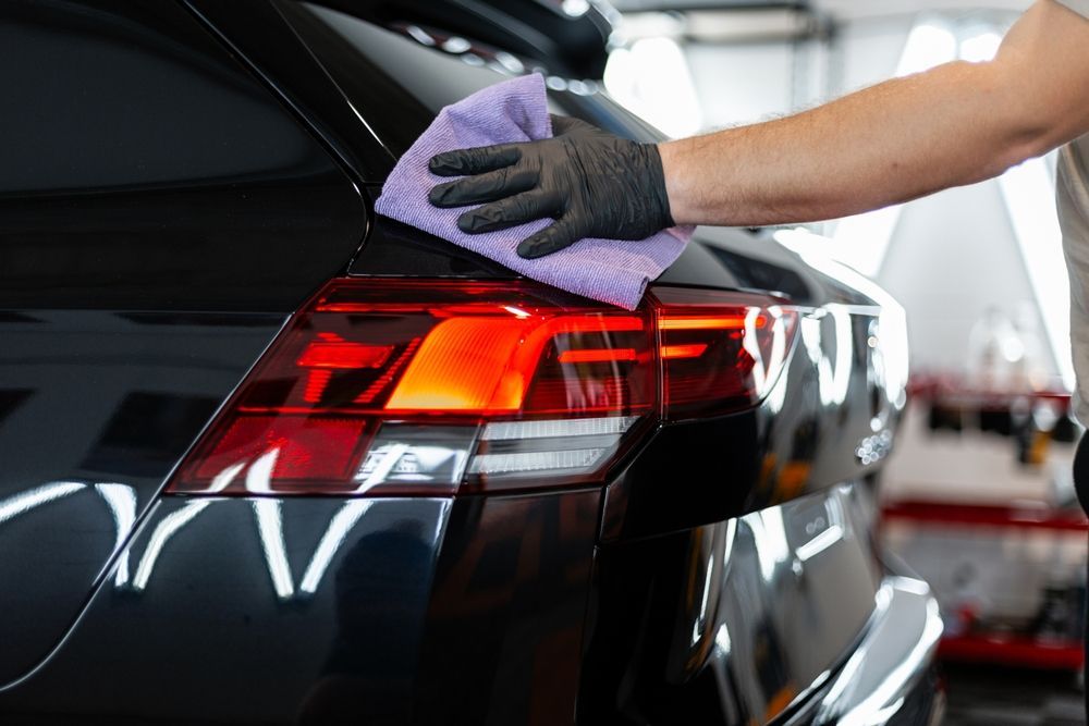 Person in black gloves wiping a car's taillight with a purple cloth in a well-lit garage.