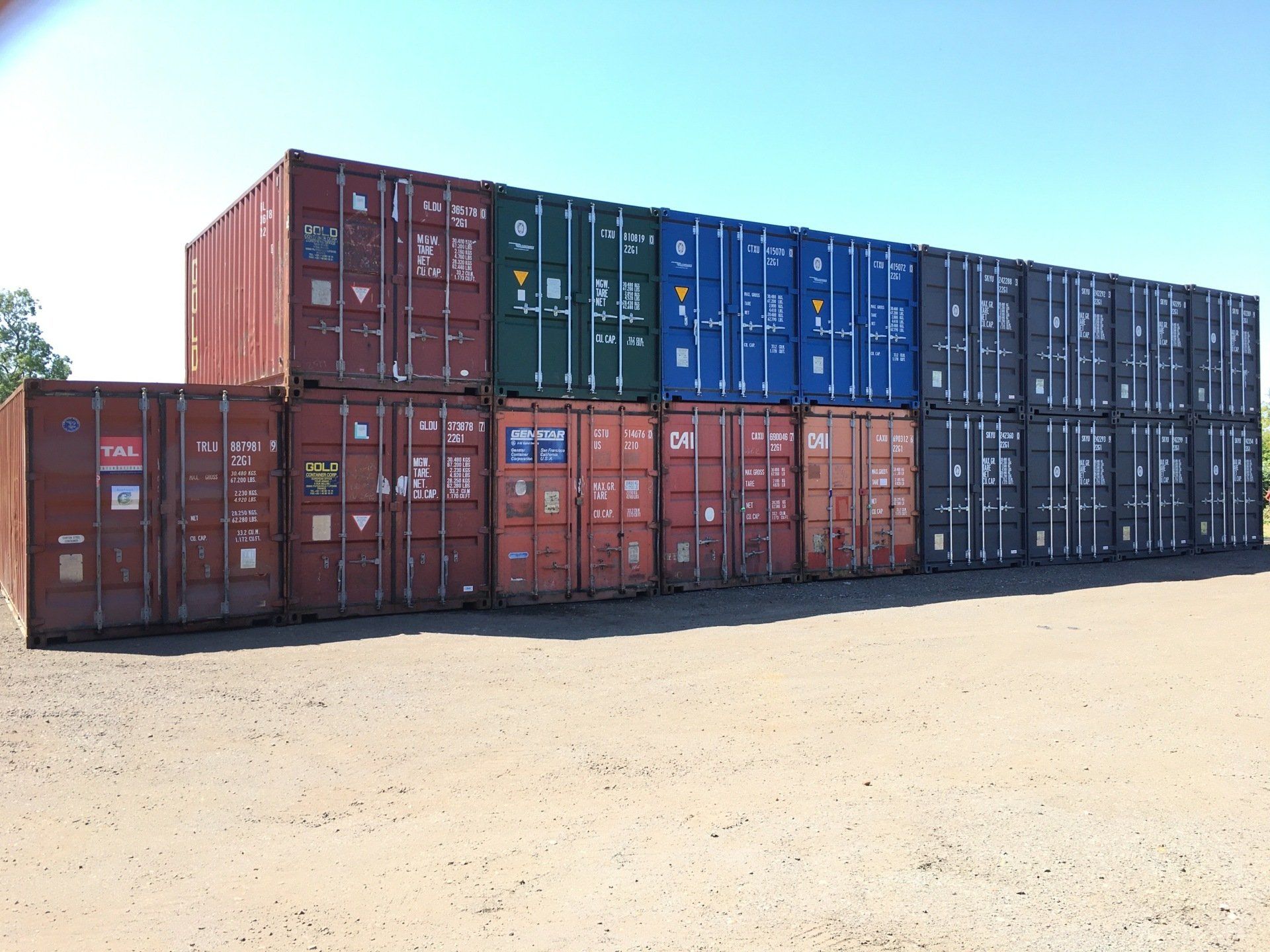 Shipping containers, various colors, stacked on a gravel lot under a clear blue sky.