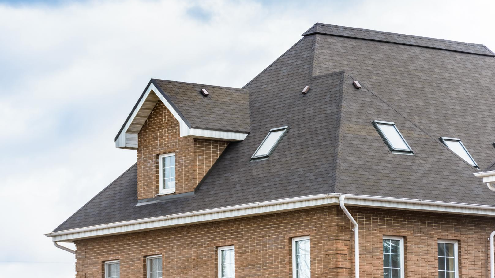 Maison en briques avec toit en bardeaux gris foncé, lucarnes, garnitures blanches et ciel clair.