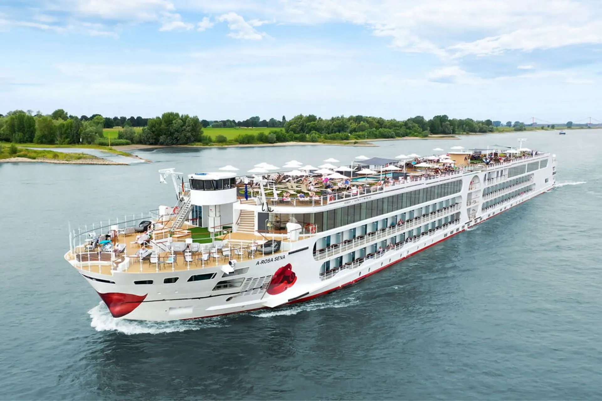 A large white A-ROSA river cruise ship with a red lip design on its bow sailing on a river past a green shoreline.