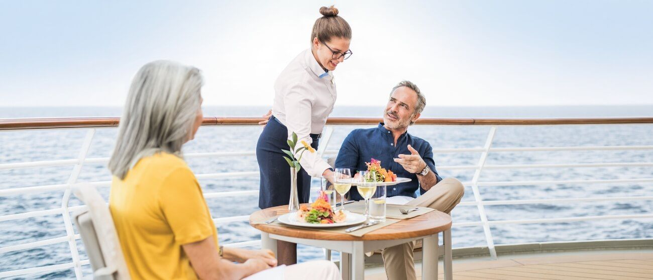 A server places a meal on a table for two on a cruise ship deck overlooking the ocean.