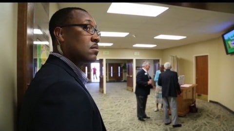 Man in suit and earpiece stands guard in a church hallway as other people gather.