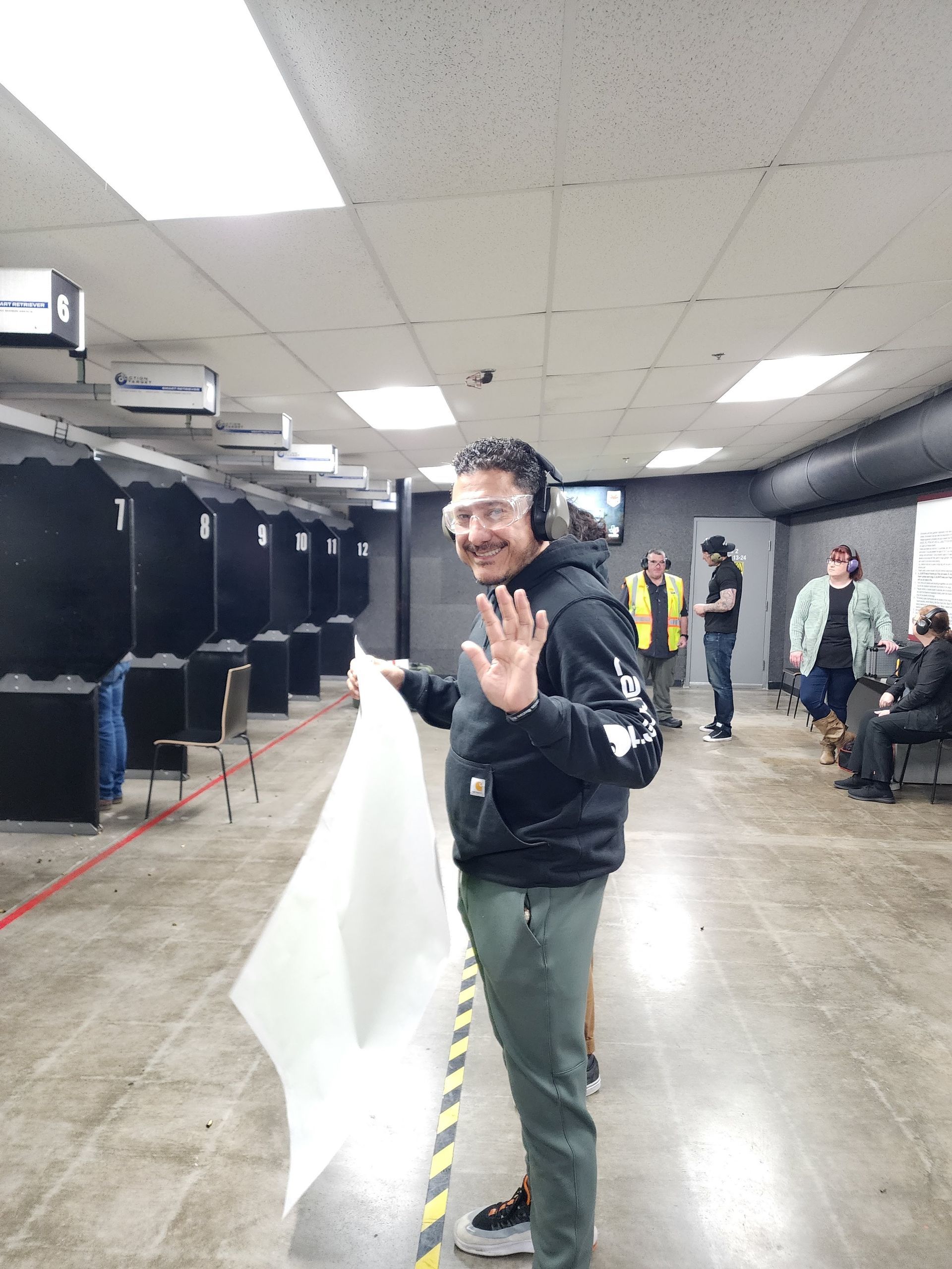 Man at a shooting range waves while holding a target. Others are in the background.