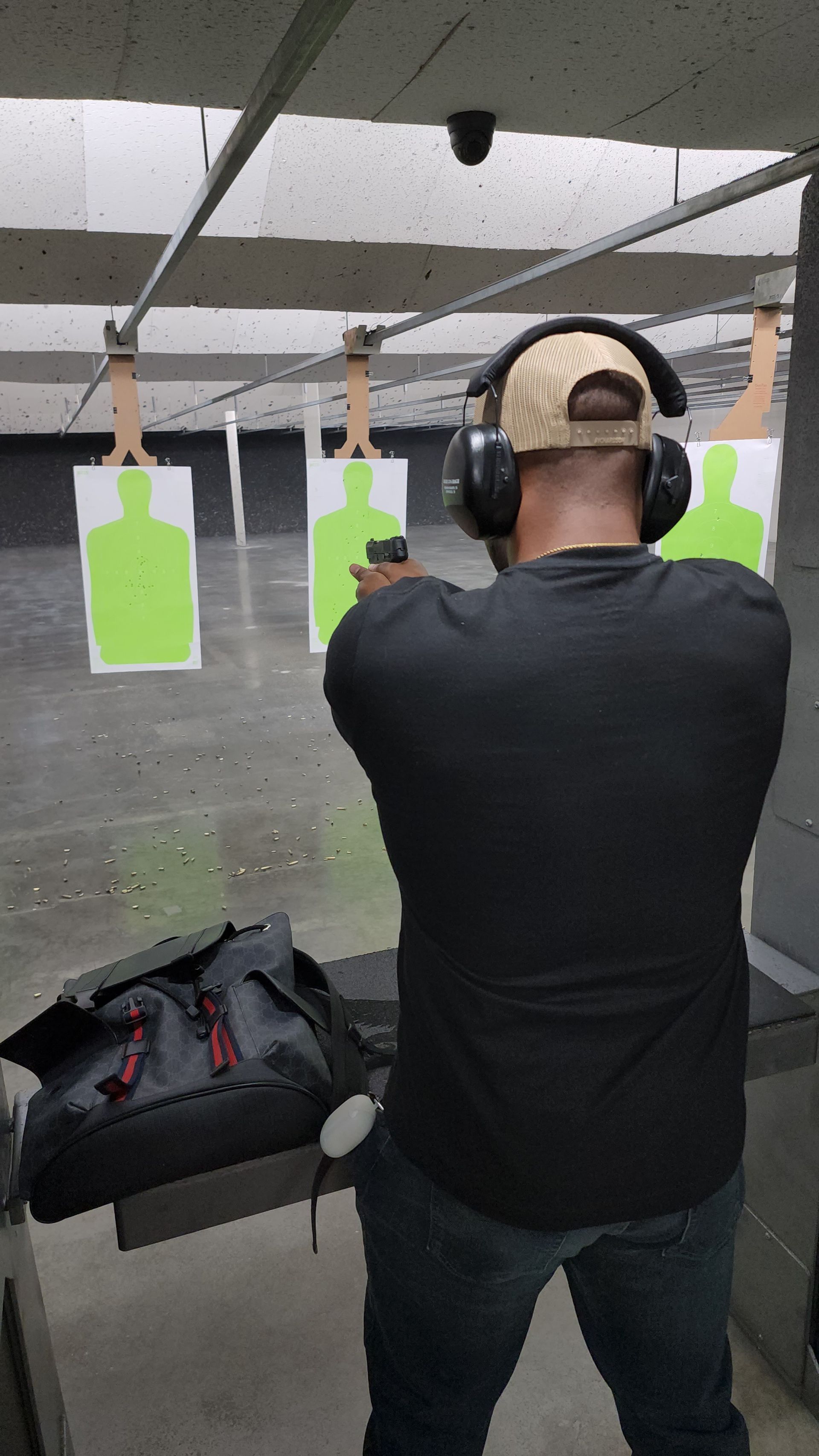 Man at a shooting range, aiming a handgun at a target; wearing earmuffs, hat, black shirt.