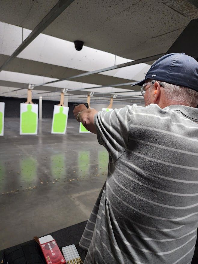 Man aiming a handgun at a target in a shooting range. Gray striped shirt, blue hat. Green and white targets in background.
