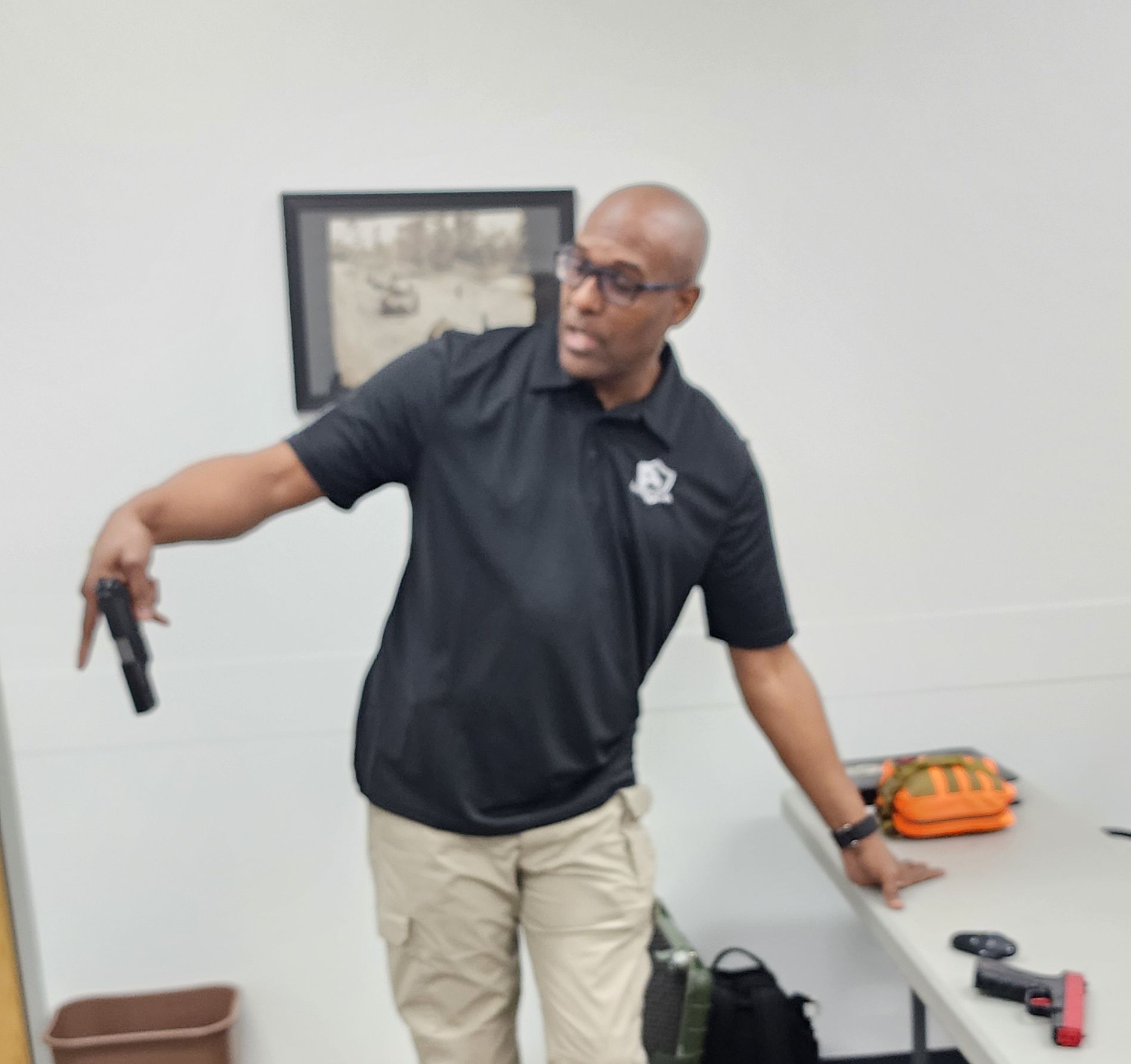 Man in black shirt, tan pants demonstrates handgun use in classroom.