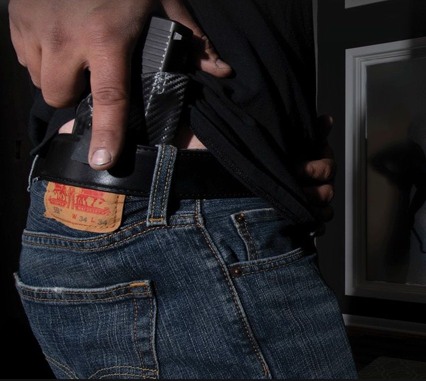 A person wearing jeans, black shirt, drawing a handgun from the waistband.